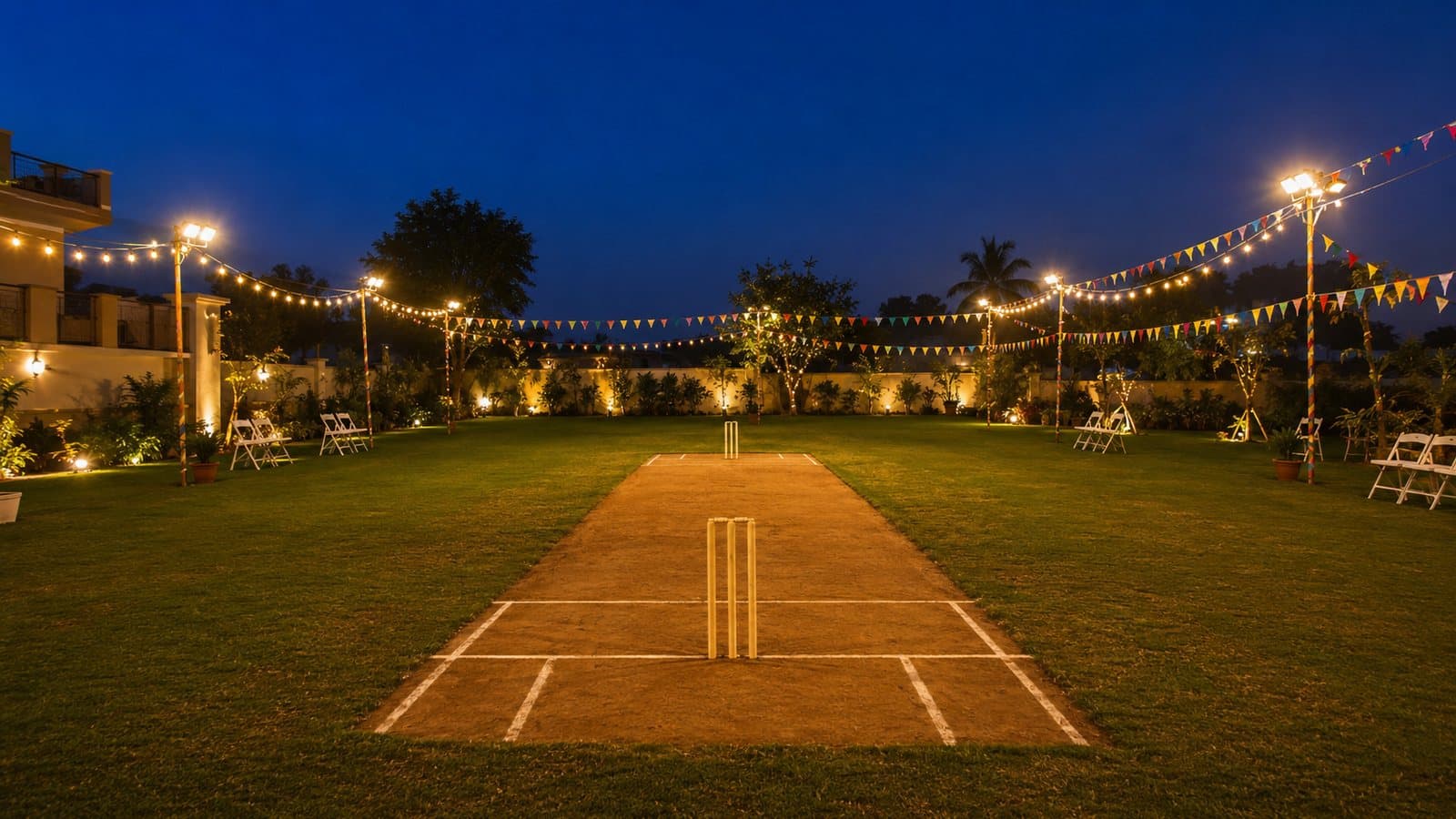 A decorated home lawn at dusk under strings of floodlit fairy lights, with a cricket pitch marked in the middle